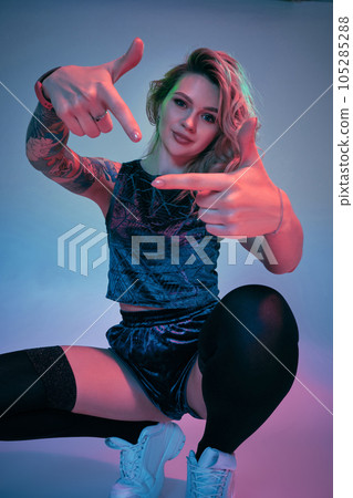 Young fitness blonde woman wearing blue velour booty shorts squats in front of camera, looking at camera, pink and blue background Young fitness blonde woman wearing blue velour booty shorts squats in front of camera, looking at camera, pink and blue background 105285288