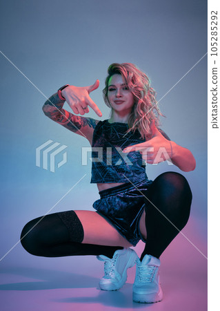 Young fitness blonde woman wearing blue velour booty shorts squats in front of camera, looking at camera, pink and blue background 105285292