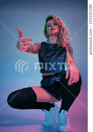 Young fitness blonde woman wearing blue velour booty shorts squats in front of camera, looking at camera, pink and blue background Young fitness blonde woman wearing blue velour booty shorts squats in front of camera, looking at camera, pink and blue background 105285296