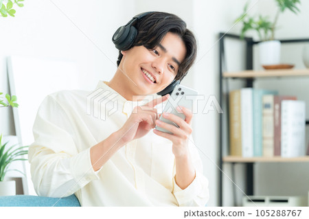 一個年輕人坐在客廳的沙發上,用智能手機和耳機聽音樂 一個年輕人坐在客廳的沙發上,用智能手機和耳機聽音樂 105288767