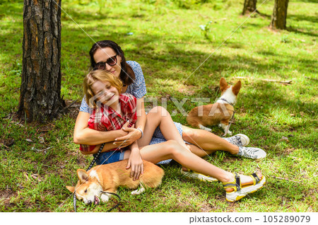 Mother hugging her little daughter resting on her sholder with closed eyes, with two corgi dogs against of green grass in the forest 105289079