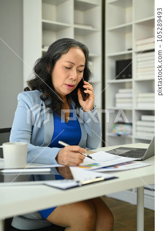 Beautiful mature businesswoman having pleased phone conversation and taking notes 105289493