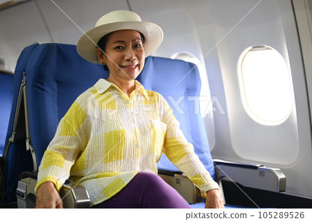Smiling middle aged woman passenger sitting in the airplane next to the window. Retirement, travel and summer vacation concept Smiling middle aged woman passenger sitting in the airplane next to the window. Retirement, travel and summer vacation concept 105289526