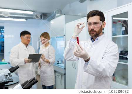 Mature experienced scientist inside laboratory researching and examining blood in test tube, man working with colleagues researchers in white medical coats. Mature experienced scientist inside laboratory researching and examining blood in test tube, man working with colleagues researchers in white medical coats. 105289682