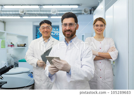 Successful team of scientists, three persons man and woman smiling and looking at the camera, portrait of a group of scientists in white medical coats inside a laboratory. Successful team of scientists, three persons man and woman smiling and looking at the camera, portrait of a group of scientists in white medical coats inside a laboratory. 105289683
