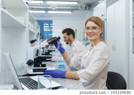 Portrait of female scientist laboratory assistant, researcher working inside medical laboratory with microscope, looking at camera and smiling while sitting, female worker in white medical coat. Portrait of female scientist laboratory assistant, researcher working inside medical laboratory with microscope, looking at camera and smiling while sitting, female worker in white medical coat. 105289696