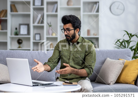 Serious focused and thoughtful Indian man talking on video call, man sitting at home in living room on sofa, using laptop for online communication with friends and colleagues. 105291571