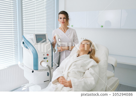 Portrait of female beautician specialist standing posing with device for photo rejuvenation in cosmetology clinic, looking at camera. Woman client receiving stimulating electric facial treatment. Portrait of female beautician specialist standing posing with device for photo rejuvenation in cosmetology clinic, looking at camera. Woman client receiving stimulating electric facial treatment. 105294086