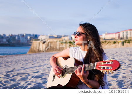 Young Latin woman play acoustic guitar on the Corunna Beach in Galicia Spain. Enjoy the travel time on summer holidays playing chill and relax 105301924