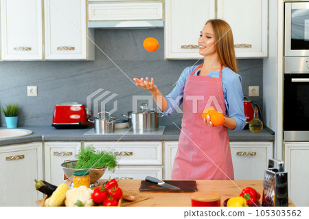 Beautiful caucasian woman in red apron holding ripe oranges while standing in kitchen Beautiful caucasian woman in red apron holding ripe oranges while standing in kitchen 105303562