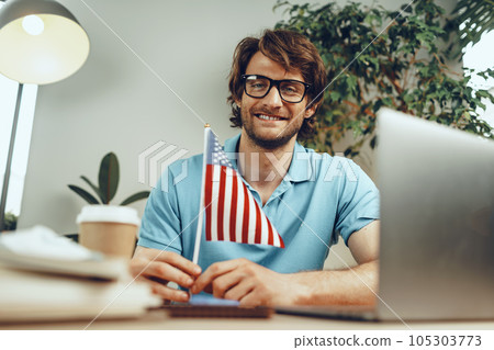 Young bearded businessman sitting at table with laptop and american flag Young bearded businessman sitting at table with laptop and american flag 105303773