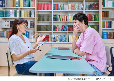 Female college counselor talking with young male student, meeting in library 105305087