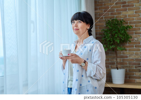 Calm beautiful mature woman with cup of coffee at home near window Calm beautiful mature woman with cup of coffee at home near window 105305130
