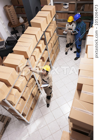 Post office storage employees managing parcels receiving and dispatching. Mail sorting center workers supervising orders audit, standing in storehouse aisle near shelves full of boxes top view Post office storage employees managing parcels receiving and dispatching. Mail sorting center workers supervising orders audit, standing in storehouse aisle near shelves full of boxes top view 105306473