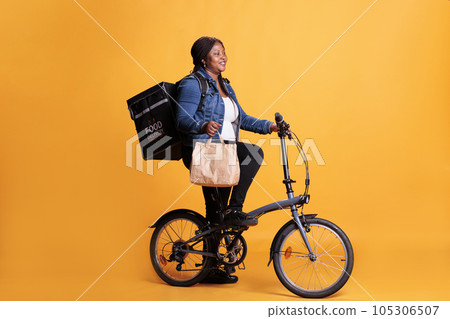 African american delivery woman holding paper bag while delivering lunch order to client using bike as transportation. Restaurant courier carrying takeout thermal backpack, takeaway service concept African american delivery woman holding paper bag while delivering lunch order to client using bike as transportation. Restaurant courier carrying takeout thermal backpack, takeaway service concept 105306507