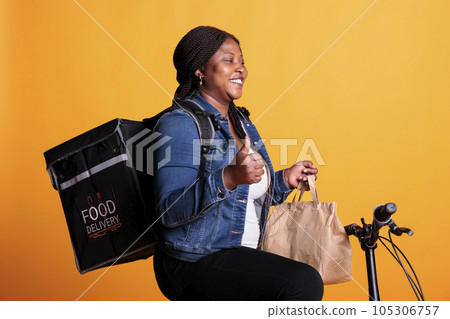 Positive courier holding paper bag full with takeaway food order delivering to customer during lunch time, worker doing thumbs up gesture at camera. Deliverywoman using bike as transportation Positive courier holding paper bag full with takeaway food order delivering to customer during lunch time, worker doing thumbs up gesture at camera. Deliverywoman using bike as transportation 105306757