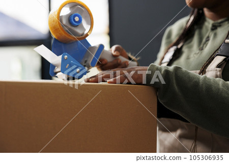 Cheerful worker putting adhesive tape on cardboard box, preparing customers packages for delivery in storehouse. Storage room manager wearing industrial overall working at online orders. Close up Cheerful worker putting adhesive tape on cardboard box, preparing customers packages for delivery in storehouse. Storage room manager wearing industrial overall working at online orders. Close up 105306935