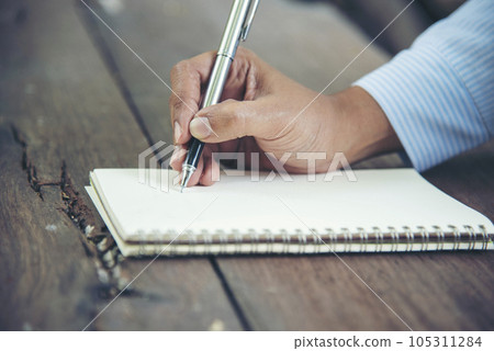 Man hands writing notebook diary with coffee cup and smartphone on wood desk. Close up man hands using pen sitting at wooden table outdoors lifestyle. Men write planner note diary office agenda Man hands writing notebook diary with coffee cup and smartphone on wood desk. Close up man hands using pen sitting at wooden table outdoors lifestyle. Men write planner note diary office agenda 105311284