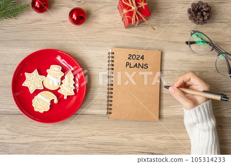 Woman hand writing 2024 PLANS on notebook with Christmas cookies on table. Xmas, Happy New Year, Goals, Resolution, To do list, and Strategy concept 105314233
