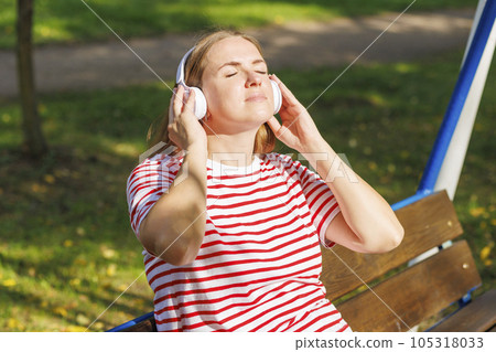A woman wearing headphones listening to music while sitting on a bench A woman wearing headphones listening to music while sitting on a bench 105318033