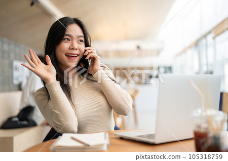 A woman is waving her hand to greet her friend while talking on the phone in a coffee shop. A woman is waving her hand to greet her friend while talking on the phone in a coffee shop. 105318759