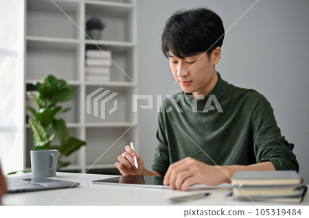 A concentrated Asian man using his digital tablet to manage his tasks at his desk in the office. A concentrated Asian man using his digital tablet to manage his tasks at his desk in the office. 105319484