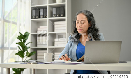 A professional and thoughtful Asian senior businesswoman focuses on her work at her desk 105319863