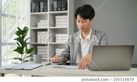 A professional Asian businessman focuses on reading business documents at his desk. A professional Asian businessman focuses on reading business documents at his desk. 105319879