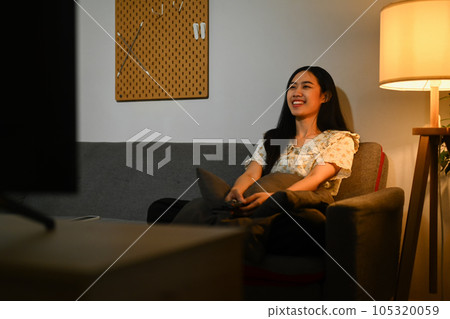 Joyful young woman watching romantic movie on TV at home in evening alone Joyful young woman watching romantic movie on TV at home in evening alone 105320059