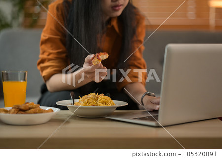 Cropped shot of young woman eating pizza and using laptop in living room. Takeaway, fast food and domestic life Cropped shot of young woman eating pizza and using laptop in living room. Takeaway, fast food and domestic life 105320091