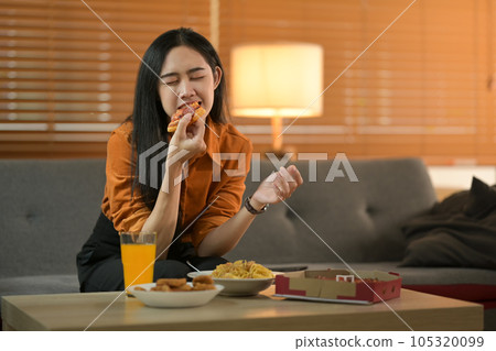 Shot of happy young Asian woman eating pizza, eating tasty lunch with closed eyes while sitting on couch Shot of happy young Asian woman eating pizza, eating tasty lunch with closed eyes while sitting on couch 105320099