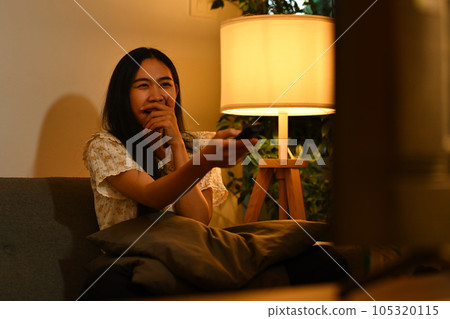 Happy young with hands clasped over mouth, excited to watch favorite movie online series at night 105320115
