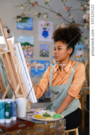 Portrait of concentrated female artist wearing apron painting picture with brush on easel in art studio 105320122