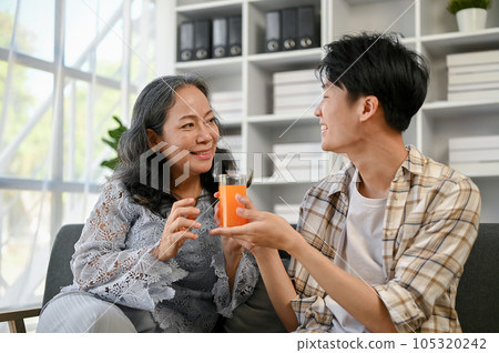 A cute and caring Asian grandson is giving a glass of orange juice to his grandmother 105320242
