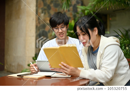 A focused Asian female college student is reading a book while her male friend is using a tablet A focused Asian female college student is reading a book while her male friend is using a tablet 105320755