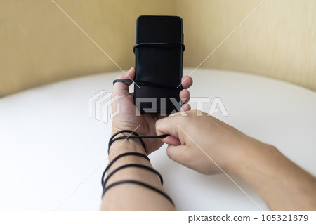 Overcoming phone addiction. Man hand is wrapped around the wire of smartphone charger. Conceptual photo of smartphone addiction. Loss of ability to live normal life. Negative impact of digitalization Overcoming phone addiction. Man hand is wrapped around the wire of smartphone charger. Conceptual photo of smartphone addiction. Loss of ability to live normal life. Negative impact of digitalization 105321879
