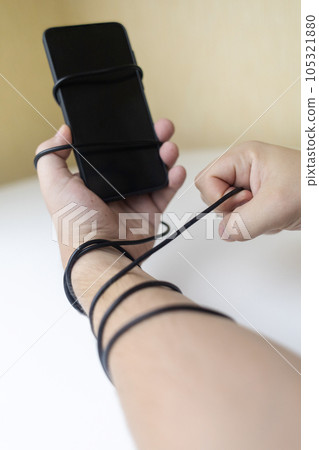 Overcoming phone addiction. Man hand is wrapped around the wire of smartphone charger. Conceptual photo of smartphone addiction. Loss of ability to live normal life. Negative impact of digitalization 105321880