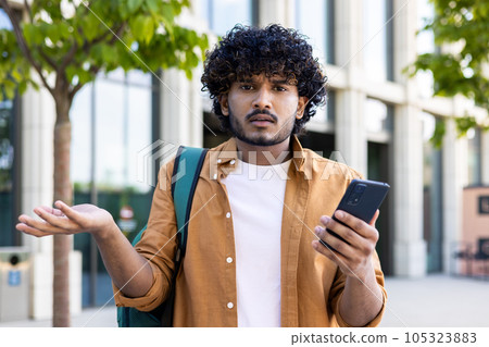 Worried young Indian man standing outside on the street, holding the phone. He spreads his hands, looks disappointedly at the camera. 105323883