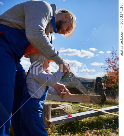 Father with toddler son building wooden frame house. Male builders using hand saw to cut boards on construction site, wearing helmet and blue overalls on sunny day. Carpentry and family concept. Father with toddler son building wooden frame house. Male builders using hand saw to cut boards on construction site, wearing helmet and blue overalls on sunny day. Carpentry and family concept. 105324136
