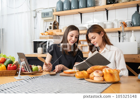 Happy two young women working cooking while her is reading a cookbook. Two young diverse lesbian women spending time together. LGBT and gender identity concept Happy two young women working cooking while her is reading a cookbook. Two young diverse lesbian women spending time together. LGBT and gender identity concept 105325148