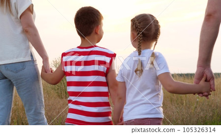 Healthy little children with parents walk joining hands across meadow lit by sunset light. Caring parents with children enjoy country nature exploration. Parents and children walk at pastureland 105326314
