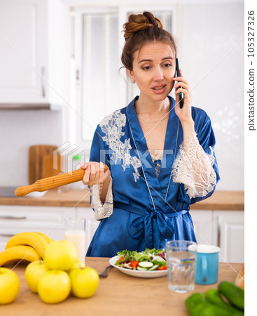 Dissatisfied woman with rolling pin in hands talking on phone in kichen 105327326