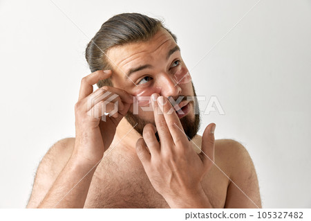 Man looking up and puts patch on area under eyes over isolated grey background. Bearded man. Man looking up and puts patch on area under eyes over isolated grey background. Bearded man. 105327482