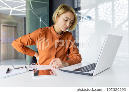 Tired and overworked woman working at workplace inside office, business woman has severe back pain, upset blonde massaging muscle, long sitting work. 105329293