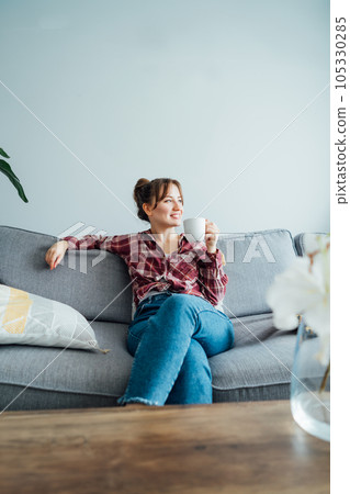 Young smiling woman sitting on sofa and looking side up while drinking coffee. Young brunette woman relaxing after housekeeping, home cleaning. Portrait of relaxed female resting at home. Vertical. 105330285