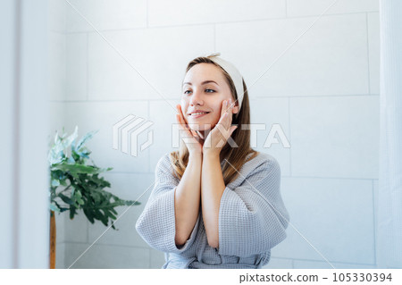 Young smiling woman applying cream mask and doing manual self massage of face in the bathroom. Natural procedures for skin care. Home Beauty treatment, face gymnastics for youth. Selective focus. 105330394