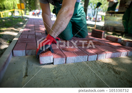 A skilled construction worker lays red paving stones on a sidewalk. 105330732