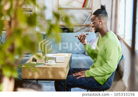 Young african man in earphones sitting table, talking on mobile phone, working remotely at home. Distance work and learning 105333156