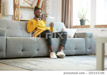 Young african an, freelancer sitting on sofa at home and having online business meeting via ablett, meeting with colleagues 105333196