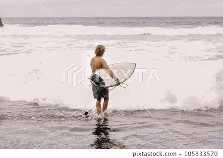 Back view of young fit surfer man with surfboard runs into ocean or sea with big waves for surfing. Concept of extreme sport, fitness, freedom, happiness, new modern life. Sports travel destination. 105333570